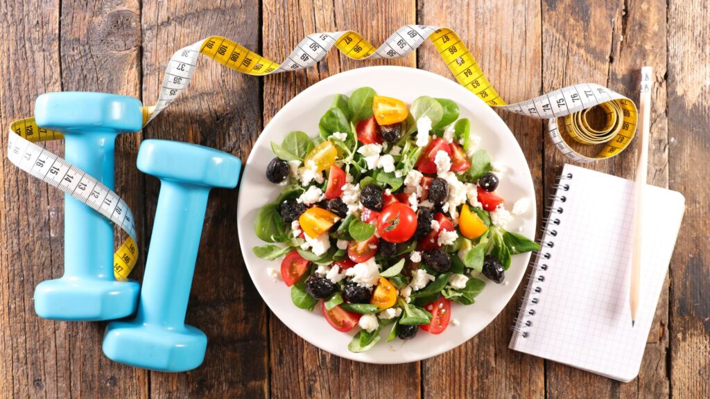 Plate of salad next to a weight, notebook, and measuring tape.